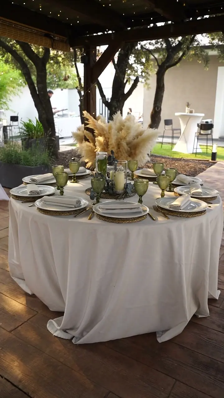 Mesa redonda de boda montada bajo una pérgola, con centro de pampas y velas