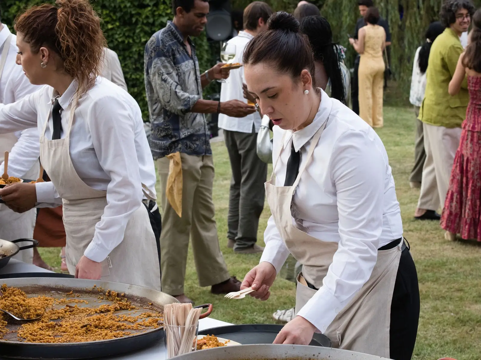 Dos cocineras de Sabor Ibérico sirven paellas en el jardín de una boda, con invitados al fondo