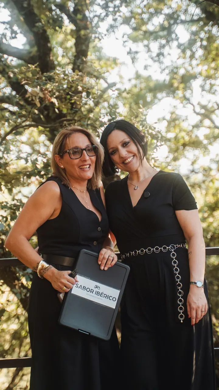 Dos responsables de un catering, vestidas de negro, sonríen en un evento al aire libre con una carpeta de Sabor Ibérico